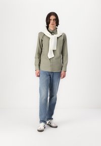 Green button-up shirt with long sleeves, worn over light blue jeans. A white scarf is draped around the shoulders. Casual shoes visible at the feet.