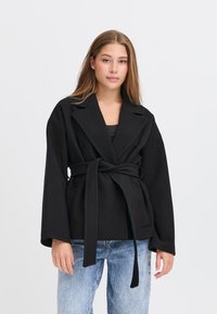 Giovane donna che indossa un cappotto nero con cintura, dalle ampie rever e jeans azzurri, in piedi davanti a uno sfondo bianco semplice.