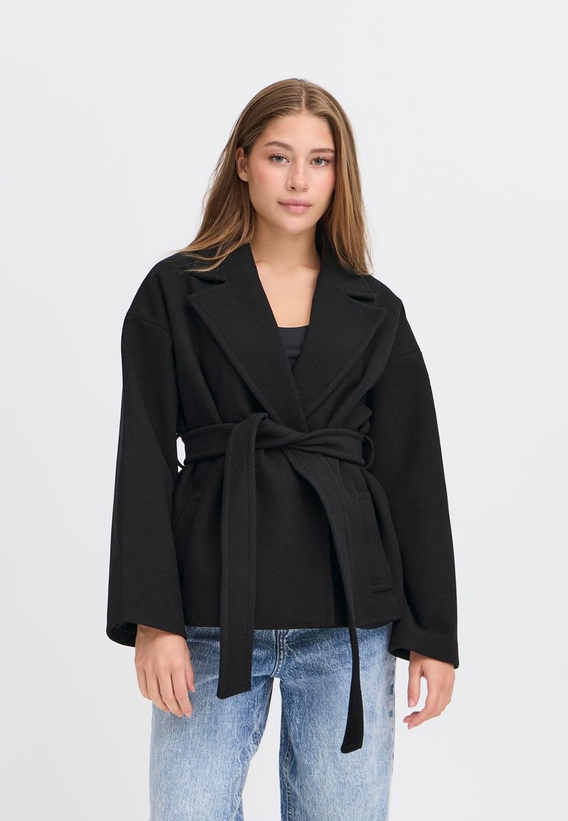 Giovane donna che indossa un cappotto nero con cintura, dalle ampie rever e jeans azzurri, in piedi davanti a uno sfondo bianco semplice.