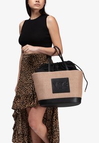 Bolsa de tote de paja trenzada con una parte superior de tela negra con cordón, que presenta una base de cuero negro y un parche con el logotipo. Mangos oscuros complementan el diseño.