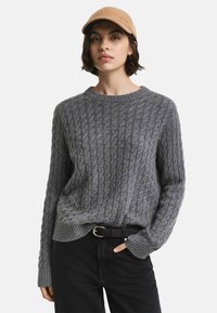 Grauer Strickpullover mit strukturiertem Kabelmuster, Rundhalsausschnitt und gerippten Bündchen, kombiniert mit schwarzen Jeans und einer beigen Mütze.