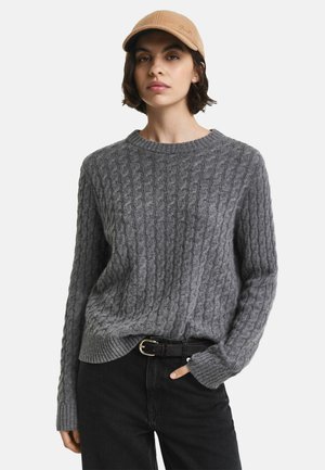 Grauer Strickpullover mit strukturiertem Kabelmuster, Rundhalsausschnitt und gerippten Bündchen, kombiniert mit schwarzen Jeans und einer beigen Mütze.
