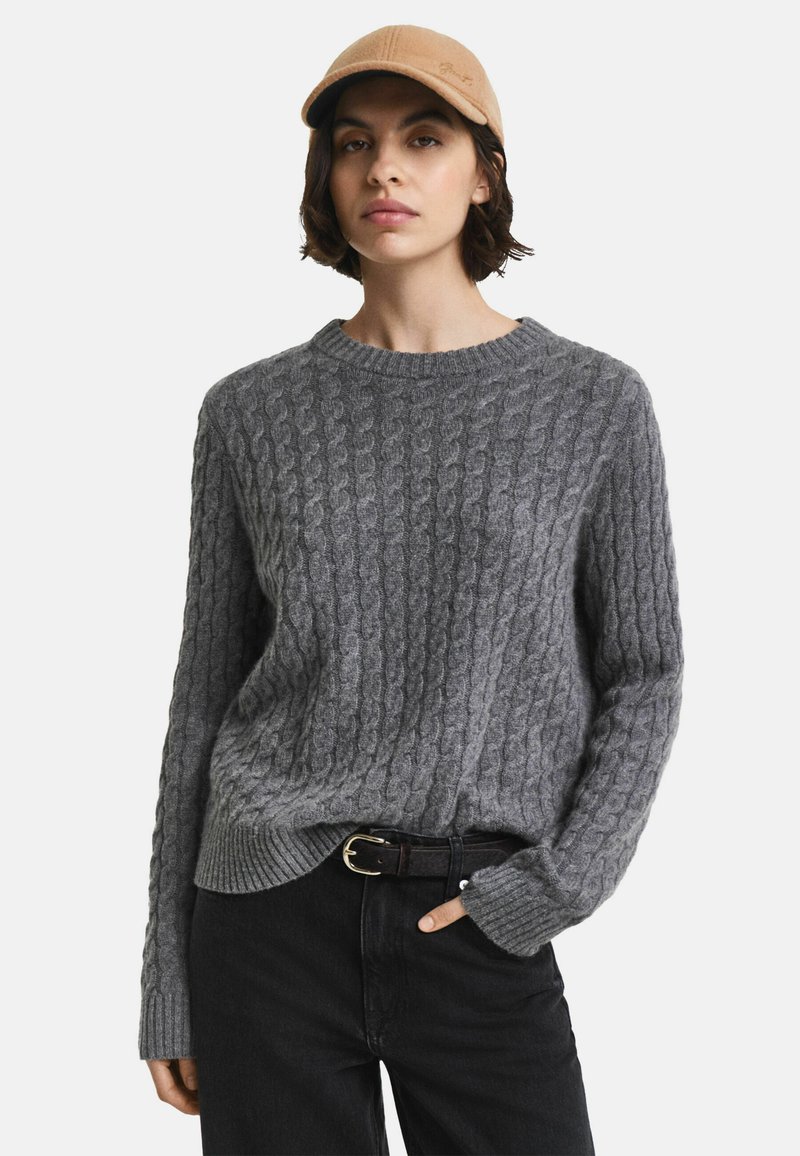 Grauer Strickpullover mit strukturiertem Kabelmuster, Rundhalsausschnitt und gerippten Bündchen, kombiniert mit schwarzen Jeans und einer beigen Mütze.