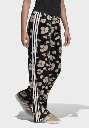 Person trägt schwarze Hose mit beigem Blumenmuster und weißen Seitenstreifen, kombiniert mit beigen Sneakers mit weißen Streifen.