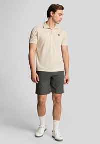 Beige polo skjorte med korte ermer og en gul logo, kombinert med mørkegrønn shorts og hvite ankelsokker med hvite joggesko.
