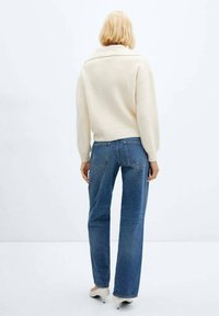 Pull en maille ivoire avec col roulé, porté avec un jean bleu coupe décontractée. Texture lisse, poignets côtelés et détail de design au dos.