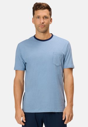 Mann mit kurzen braunen Haaren, der ein hellblaues kurzärmeliges T-Shirt mit Brusttasche und dunkelblauem Kragen auf einfarbigem Hintergrund trägt.