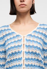 Γυναίκα που φοράει μπλε και λευκή ριγέ πλεκτή ζακέτα με στρογγυλά λευκά κουμπιά και V-λαιμόκοψη, εμφανιζόμενη από το πιγούνι μέχρι το στήθος.