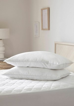 Zwei weiße Kissen auf einer gesteppte Matratze gestapelt in einem minimalen Schlafzimmer mit einer Lampe und gerahmter Wandkunst.