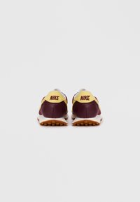 Nike sportbačiai, pagaminti iš raudonai rudos ir geltonos medžiagos, su tinkliniais paviršiais, paminkštintu padu ir ryškiu prekės ženklo logotipu ant kulno.