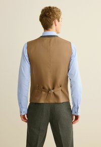 Brun vest med en glat tekstur, justerbar rygstrop med metalhardware, båret over en lyseblå skjorte, parret med grå mønstrede bukser.