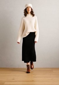 Una donna indossa un maglione a coste color crema e una gonna nera midi, abbinati a stivali marroni con lacci. Ha un berretto color crema abbinato.