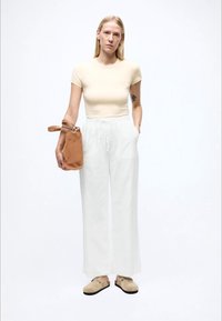 Béžové priliehavé tričko, biele široké nohavice s vreckami a svetlohnedá taška. Nohy v béžových nazúvacích topánkach, minimalistický dizajn, jednoduché pozadie.