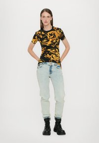 Svart t-skjorte med gult dekorativt mønster, korte ermer; kombinert med lyseblå, avslappet passform jeans og svarte snørebånd ankel støvler.