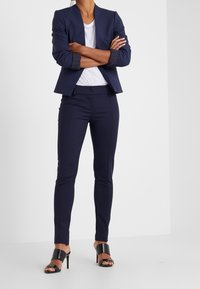 Femme debout les bras croisés, portant un blazer bleu marine, un t-shirt blanc, un pantalon bleu marine assorti et des sandales à talons noirs, sur fond blanc.