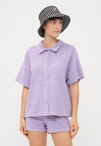 Camicia corta a maniche corte a righe viola e bianche abbinata a pantaloni corti coordinati. Un cappello a secchiello a scacchi bianco e nero completa l'outfit.