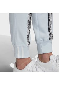 Ljust blå idrottsbyxor med svart Adidas-logotyp längs sidorna. Ribbad mudd och tre vita ränder vid anklarna. Vita sneakers synliga.