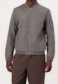 Šedá bomber bunda se zipem vpředu, bočními kapsami a kapsou na rukávu. Má ribovaný límec a manžety, vyrobená z hladkého, lehkého materiálu.