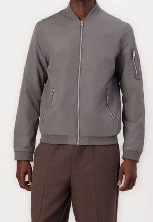 Harmaa pommilentäjän takki, jossa on vetoketju edessä, sivutaskut ja hihnatasku. Varustettu ribbineuloksella kauluksessa ja hihansuissa, valmistettu sileästä, kevyestä materiaalista.