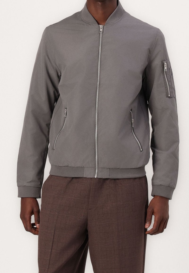 Šedá bomber bunda se zipem vpředu, bočními kapsami a kapsou na rukávu. Má ribovaný límec a manžety, vyrobená z hladkého, lehkého materiálu.