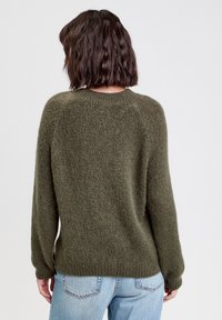 Pull en tricot vert olive avec une coupe décontractée, col rond et poignets côtelés. La texture est douce et moelleuse, associé à un jean en denim bleu clair.