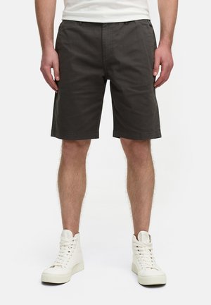Pantalones cortos gris oscuro de algodón, con un diseño de frente plano, trabillas para cinturón y bolsillos laterales. Combinados con zapatillas altas blancas.