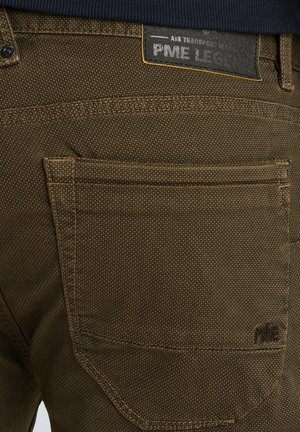 Pantalones de tela marrón con tejido texturizado, que cuentan con un bolsillo trasero y una etiqueta negra con la marca "PME Legend", acentuada con costuras amarillas.