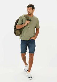 Oliven grøn t-shirt med korteærmer, parret med opviklede denimshorts og hvide sneakers med marineblå detaljer. Brun rygsæk med lommer.