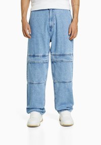 Man som bär ljusblå denimcargo-byxor och vita sneakers, stående med avslappnade händer vid sidorna på vit bakgrund.