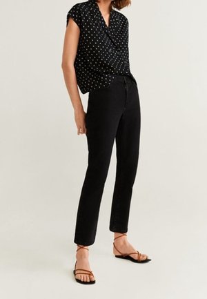 Femme portant un jean noir droit, un chemisier noir à manches courtes avec des pois blancs, et des sandales plates marron à lanières, debout devant un fond uni.