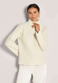Vrouw in een crèmekleurige kabelgebreide coltrui en witte broek, met gouden creolen, poseert tegen een effen lichte achtergrond.