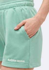 Κοντινό πλάνο ενός χεριού που ακουμπά στην τσέπη ενός σορτς Björn Borg σε μέντα πράσινο χρώμα με ελαστική μέση, πάνω σε λευκό φόντο.