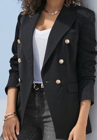 Černý dvouřadový blejzr z hladkého materiálu, zdobený zlatými knoflíky, přiléhavého st�řihu a s vyhrnutými rukávy, v kombinaci s bílým topem a džíny.