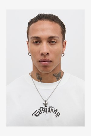 Fehér T-shirt fekete "Ed Hardy" logóval, ezüstlánc nyaklánc szívalakú medállal, feltűnő rózsa és pillangó tetoválások a nyakon.