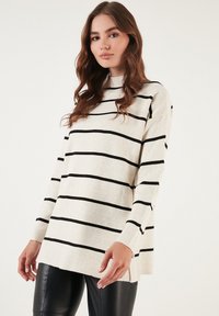 Maglione crema con righe orizzontali nere, scollo rotondo e maniche lunghe. Caratterizzato da fessure laterali e una vestibilità morbida.