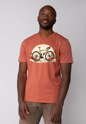 Roestkleurig katoenen t-shirt met een fietsafbeelding tegen een zonsondergang achtergrond, met palmbomen aan beide zijden geïllustreerd.