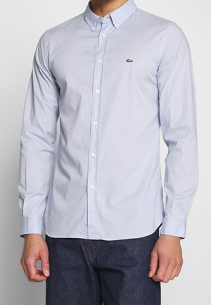 Man draagt een lichtblauw, knoopsluitend overhemd met lange mouwen, met een klein groen krokodillenlogo op de borst en donkerblauwe jeans.