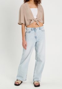 Beiges, bauchfreies Top mit Knoten vorn, weißes Untertanktop, hellblaue Jeans mit weitem Bein und beigen Sandalen mit dekorativen Akzenten.