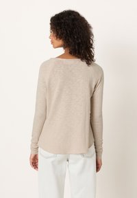 Moteris su garbanotais plaukais, vilkinti smėlio spalvos ilgomis rankovėmis palaidinę ir šviesios spalvos džinsus, matoma iš nugaros priešais paprastą foną.