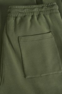 Spodnie dresowe w oliwkowym zielonym kolorze, z elastycznym pasem i tylną kieszenią z naszywką. Gładka faktura, minimalistyczny design i szczegółowe przeszycia.