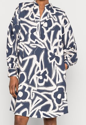 Robe à manches longues avec un motif floral abstrait bleu marine et blanc portée par une personne avec la main dans la poche.