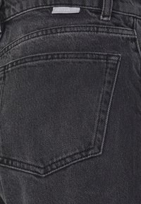 Poche arrière d'un jean en denim noir avec des coutures apparentes et une petite étiquette portant la mention "Boyish" au-dessus de la poche.