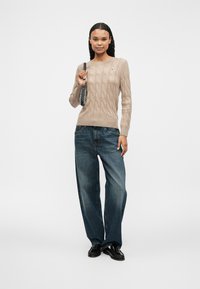 Pull en maille beige à manches longues, associé à un jean bleu décontracté et des chaussures noires. Accessoirisé avec un petit sac à main.