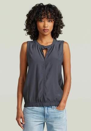 Donna con capelli ricci che indossa un top senza maniche grigio scuro con scollo a goccia e jeans azzurri, una mano in tasca.