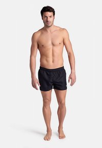 Svarta badshorts med elastisk midja, tillverkade i lätt tyg, med en logotyp på sidan, båda av en stående manlig modell.