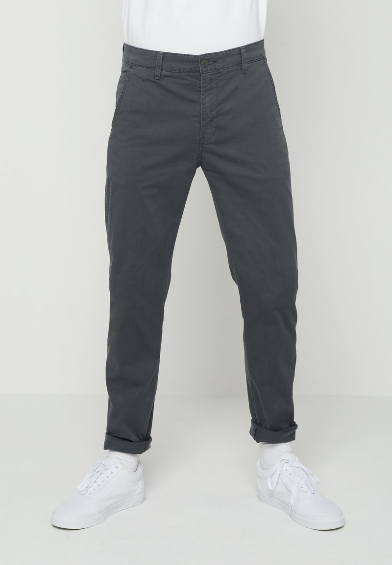 Homme portant un chino gris foncé retroussé et des baskets blanches avec des chaussettes blanches, debout devant un fond clair uni.