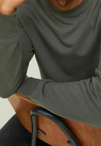 Sudadera verde oscura con puños acanalados, que presenta un sencillo cuello redondo. La tela parece suave y texturizada. Una mano reposa sobre una silla de madera.