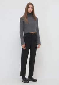 Giovane donna in piedi che indossa un maglione a collo alto grigio a coste, pantaloni neri a vita alta su misura e mocassini neri, contro uno sfondo bianco uniforme.