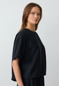 Top nero, largo, a maniche corte con scollatura rotonda. Presenta una texture morbida e liscia e un design accorciato, indossato sopra un tessuto coordinato.
