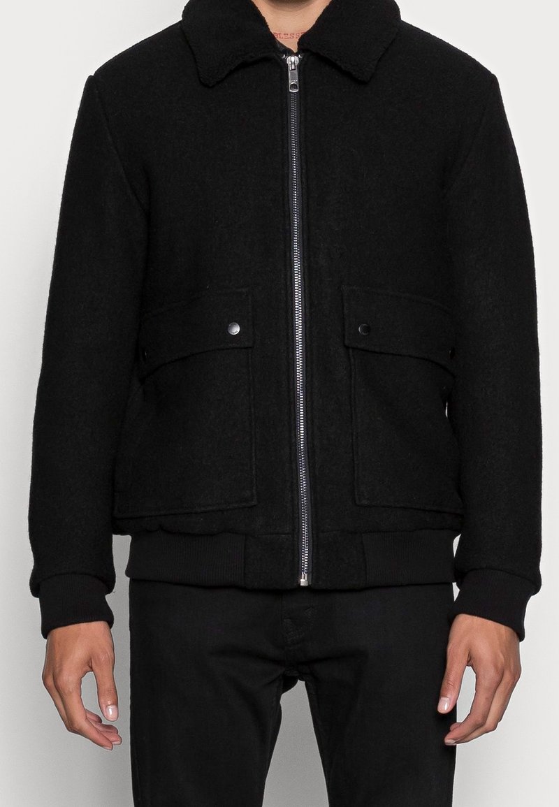 Homme portant une veste noire zippée avec col et deux poches avant à rabat avec fermeture par boutons.
